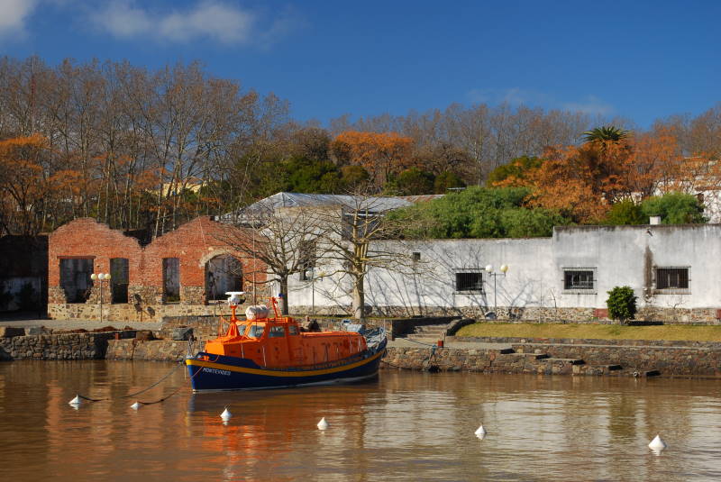 Orange and blue boat moored in Colonia del Sacramento's harbor with colonial brick buildings and autumn trees in the background