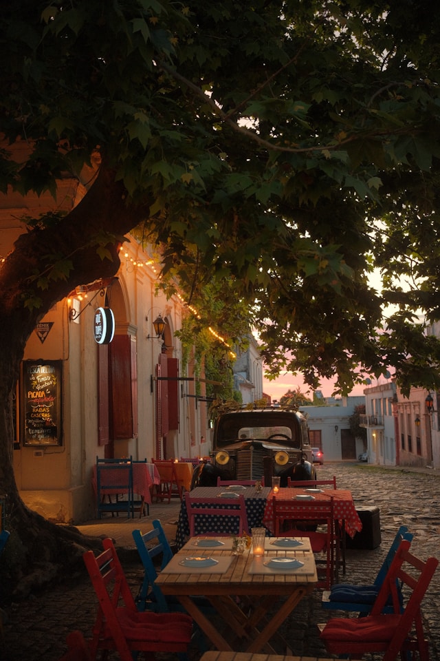 Outdoor café shaded by a large tree with a vintage car parked on the street in Colonia del Sacramento
