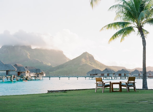 Two wooden lounge chairs on a lawn facing a lagoon lined with overwater huts in Bora Bora French Polynesia