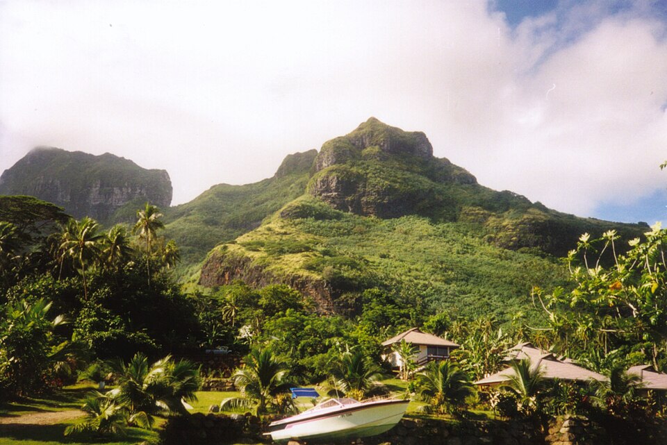 A green volcanic peak rising above palm trees and a small boat on shore