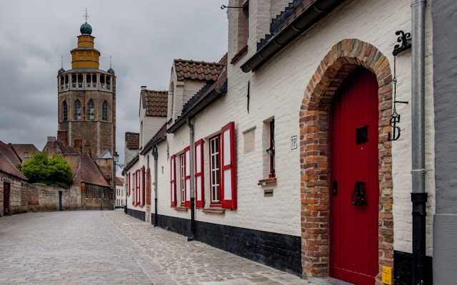 Whitewashed brick street with a red door and distant bell tower in Bruges Belgium