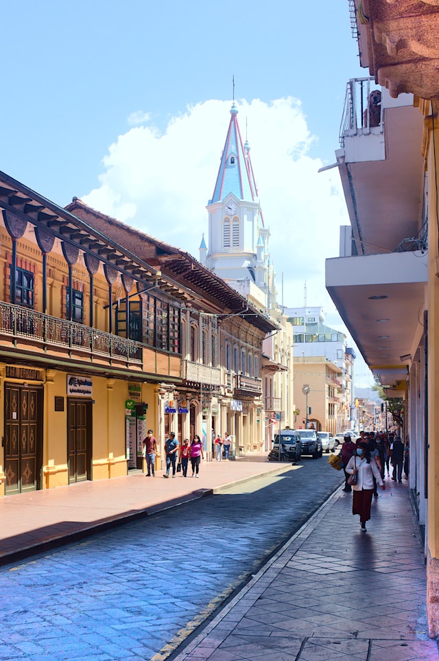 A quiet colonial street lined with balconies beneath a blue spired clock tower in Cuenca Ecuador