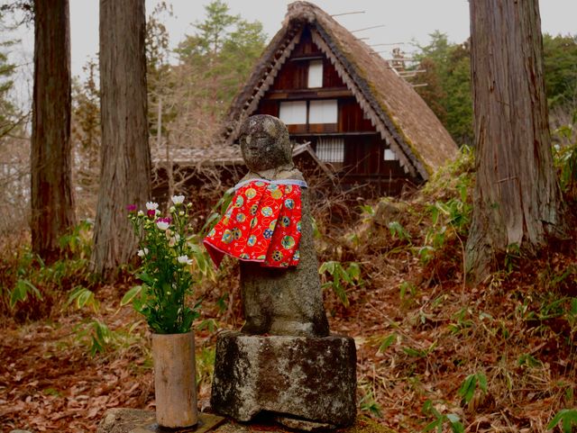 Stone jizo statue wearing a red bib near a thatched house in Takayama