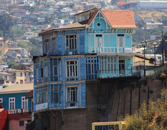 A weathered blue wooden house with bay windows standing above a retaining wall in Valparaiso Chile